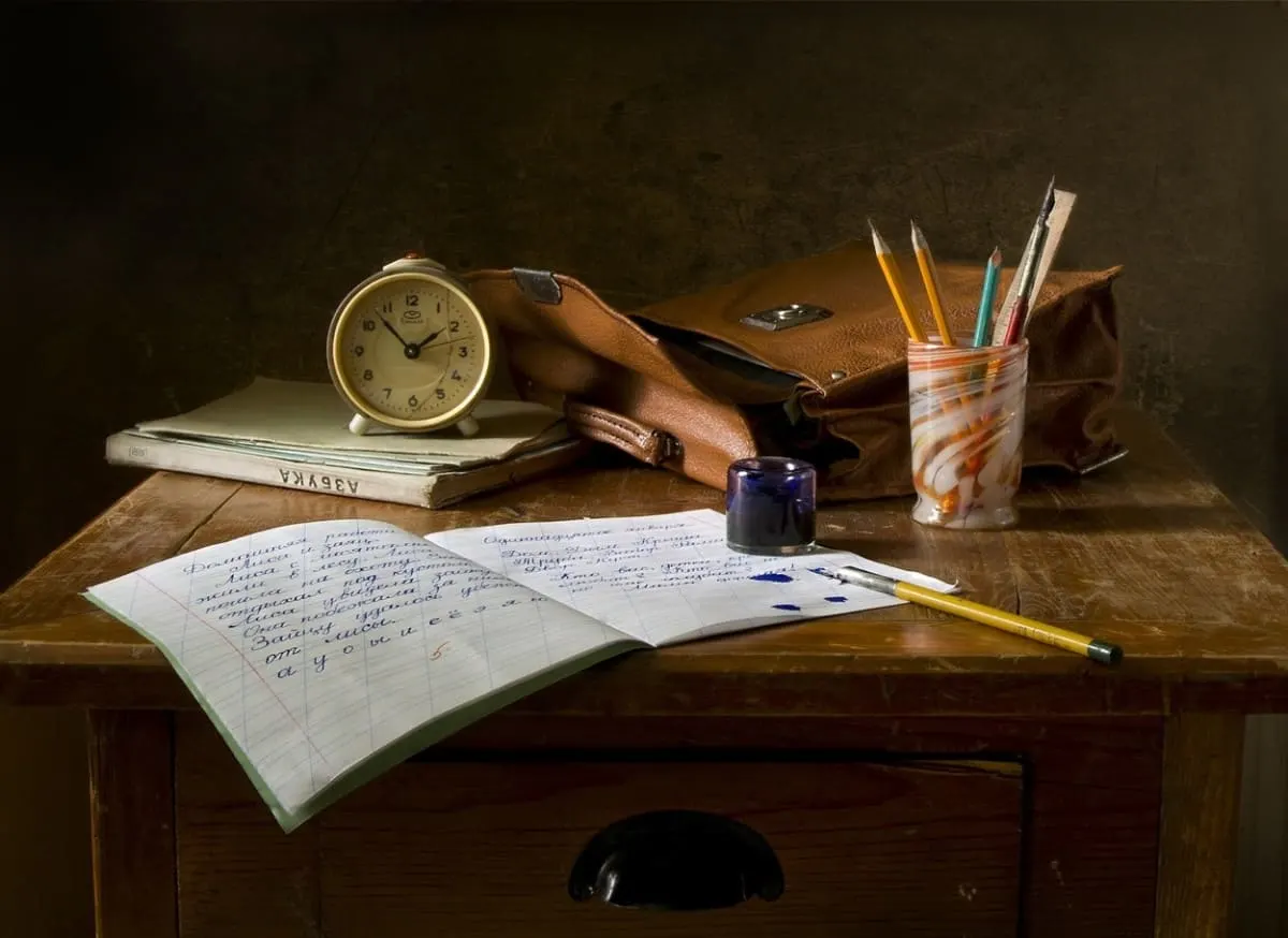 A picture of a notebook, pen, a backpack, an alarm clock with some books in a table.