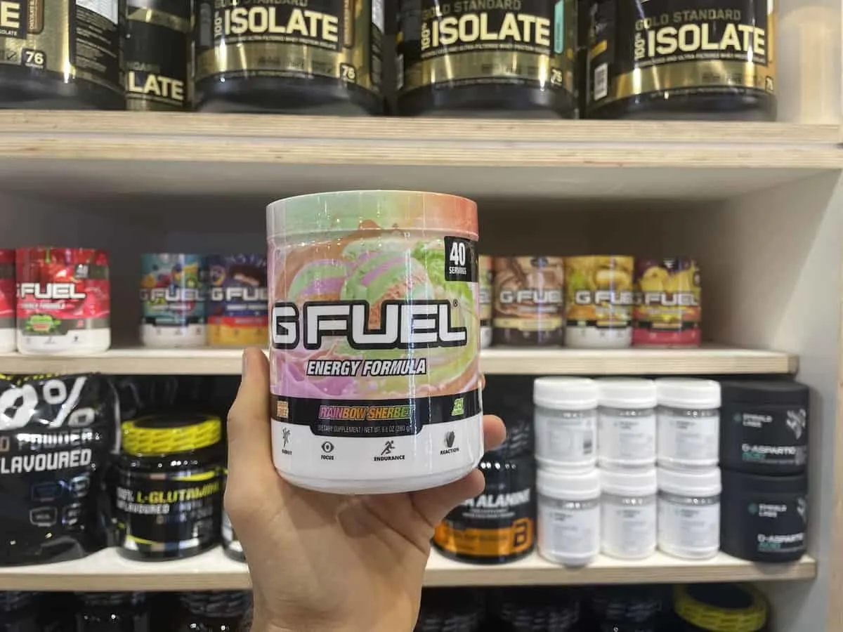 G Fuel Rainbow Sherbet energy tub held in hand with different flavors of G Fuel in background