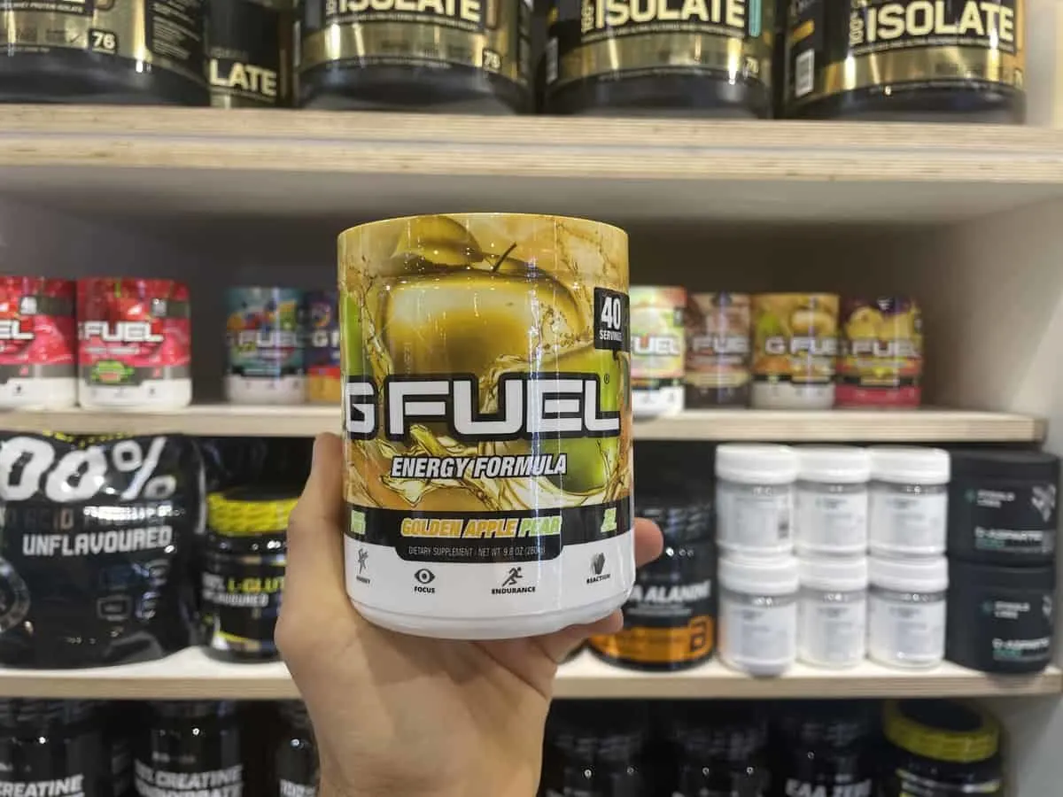 G Fuel Golden Apple Pear energy tub held in hand with different flavors of G Fuel in background