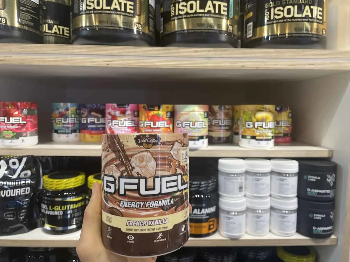 G Fuel French Vanilla energy tub held in hand with different flavors of G Fuel and supplements in background
