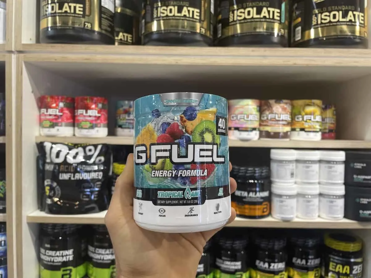 G Fuel Tropical Rain energy tub held in hand with different flavors of G Fuel and supplements in background