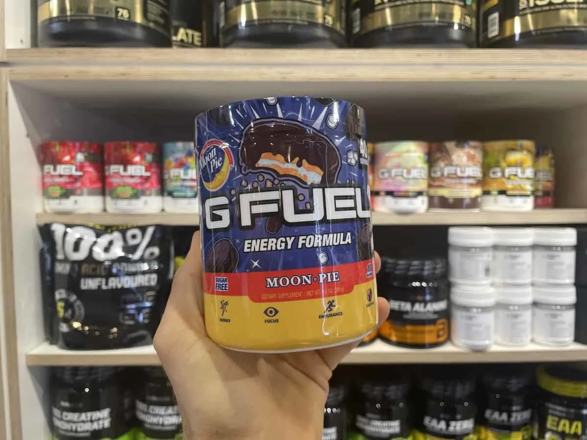G Fuel Moon Pie energy tub held in hand with different flavors of G Fuel and supplements in background